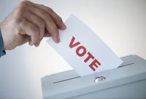 Image of a hand placing a paper with the words "vote" on it into a ballot box. 