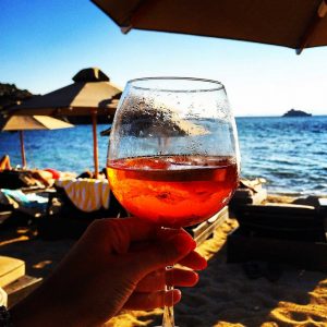 Image of a hand holding an Aperol spritz in front of a beach scene in celebration