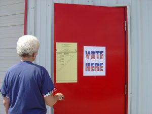 Image of a person wearing a blue shirt opening a red door. The red door has a sign that says: "Vote Here"