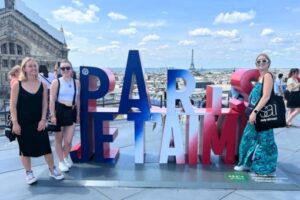 Students stand on a rooftop overlooking a city with a view of the Eiffel Tower and large letters that say "Paris, Je t'aime"